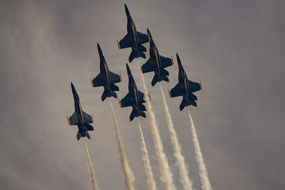 Six military jet aircraft are flying in a tight diamond formation against an overcast gray sky. Each aircraft is trailing white smoke from its engines, creating parallel contrail streams below the formation. The aircraft display dark blue and yellow color schemes consistent with a precision aerobatic demonstration team.