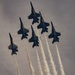 Six military jet aircraft are flying in a tight diamond formation against an overcast gray sky. Each aircraft is trailing white smoke from its engines, creating parallel contrail streams below the formation. The aircraft display dark blue and yellow color schemes consistent with a precision aerobatic demonstration team.