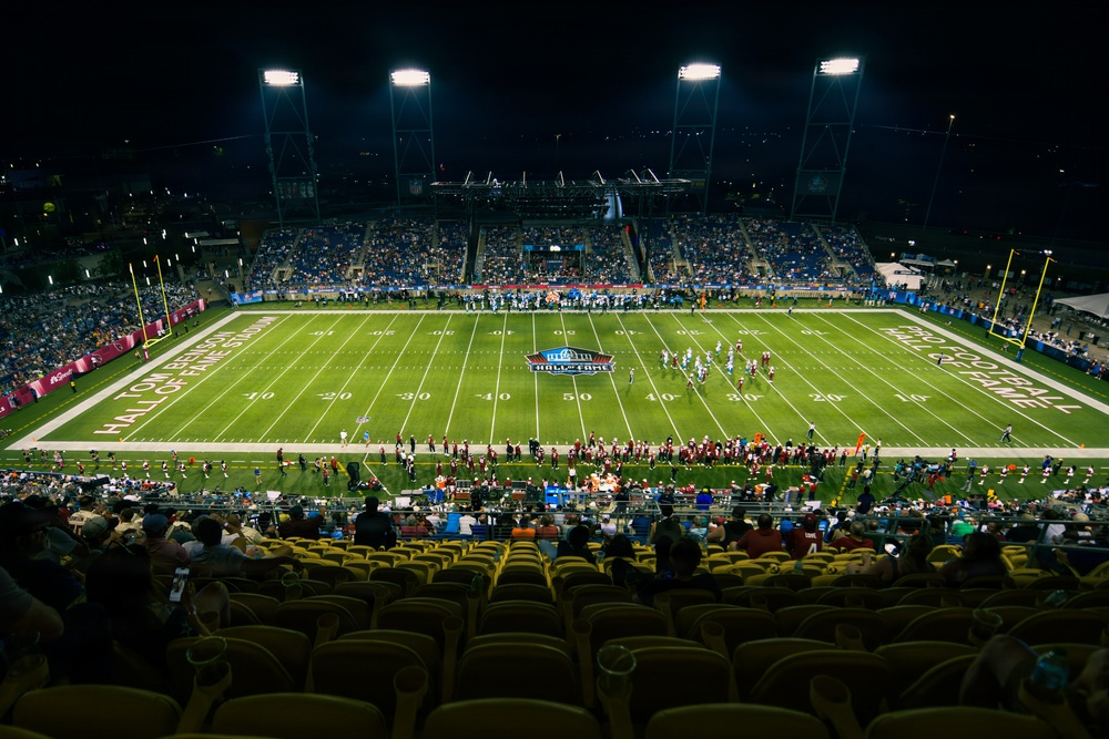 A night game at Tom Benson Hall of Fame Stadium, home of the NFL Hall of Fame Game, photographed from an elevated vantage point in the upper seating section. The stadium is lit by large floodlights, illuminating a green turf field with the Pro Football Hall of Fame logo at midfield. The stands are partially filled with spectators, and players and personnel are visible on the field and sidelines.