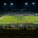 A night game at Tom Benson Hall of Fame Stadium, home of the NFL Hall of Fame Game, photographed from an elevated vantage point in the upper seating section. The stadium is lit by large floodlights, illuminating a green turf field with the Pro Football Hall of Fame logo at midfield. The stands are partially filled with spectators, and players and personnel are visible on the field and sidelines.