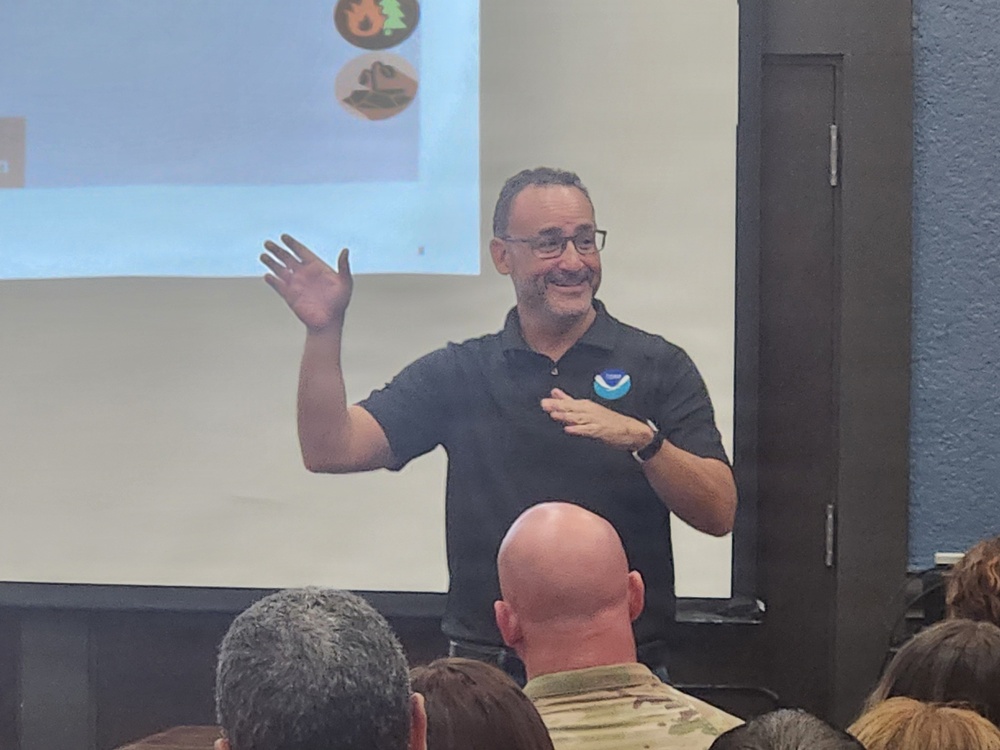 A man wearing a NOAA-logoed black polo shirt is presenting to an audience in what appears to be a conference or briefing room. He is smiling and gesturing with his right hand raised, standing in front of a projection screen displaying icons related to natural hazards. Several audience members are visible from behind in the foreground.