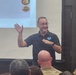 A man wearing a NOAA-logoed black polo shirt is presenting to an audience in what appears to be a conference or briefing room. He is smiling and gesturing with his right hand raised, standing in front of a projection screen displaying icons related to natural hazards. Several audience members are visible from behind in the foreground.