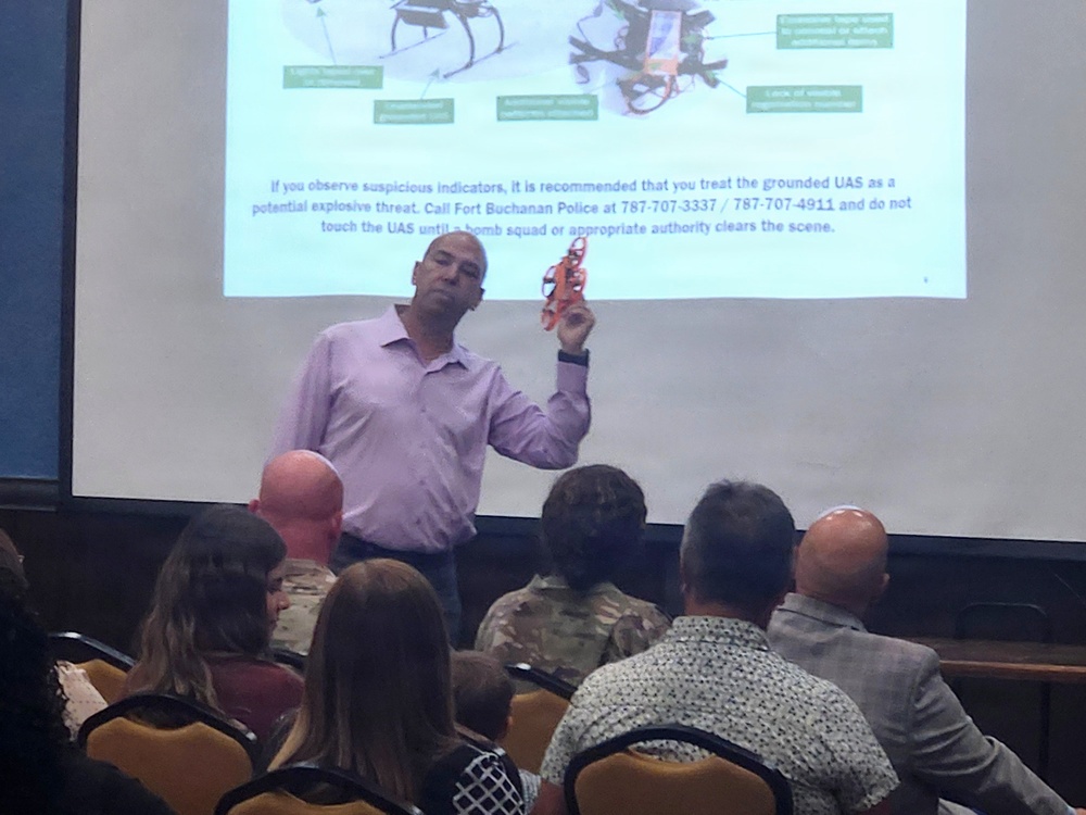 A presenter is conducting a security briefing about UAS (Unmanned Aerial Systems) threats at what appears to be Fort Buchanan. He is holding up a small orange drone as a physical example while a projected slide behind him displays information about suspicious UAS indicators and emergency contact numbers. The audience includes both military personnel in camouflage uniforms and civilians.