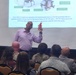 A presenter is conducting a security briefing about UAS (Unmanned Aerial Systems) threats at what appears to be Fort Buchanan. He is holding up a small orange drone as a physical example while a projected slide behind him displays information about suspicious UAS indicators and emergency contact numbers. The audience includes both military personnel in camouflage uniforms and civilians.