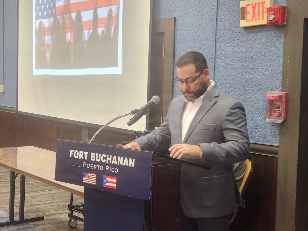 A man in a gray blazer and white shirt is speaking at a podium at Fort Buchanan, Puerto Rico. The podium displays the text 'FORT BUCHANAN PUERTO RICO' along with the American and Puerto Rican flags. Behind him is a projection screen showing an image with an American flag, and fire safety equipment is visible on the wall to the right.