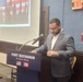 A man in a gray blazer and white shirt is speaking at a podium at Fort Buchanan, Puerto Rico. The podium displays the text 'FORT BUCHANAN PUERTO RICO' along with the American and Puerto Rican flags. Behind him is a projection screen showing an image with an American flag, and fire safety equipment is visible on the wall to the right.