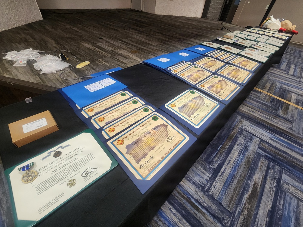 A long table displays an array of military awards and certificates arranged for a ceremony or presentation event. The documents include multiple 'Certificate of Appreciation' certificates from Fort Buchanan, Puerto Rico, along with at least one Department of the Army Army Achievement Medal citation. The setup suggests a pre-ceremony staging area where awards are organized prior to distribution.