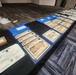 A long table displays an array of military awards and certificates arranged for a ceremony or presentation event. The documents include multiple 'Certificate of Appreciation' certificates from Fort Buchanan, Puerto Rico, along with at least one Department of the Army Army Achievement Medal citation. The setup suggests a pre-ceremony staging area where awards are organized prior to distribution.