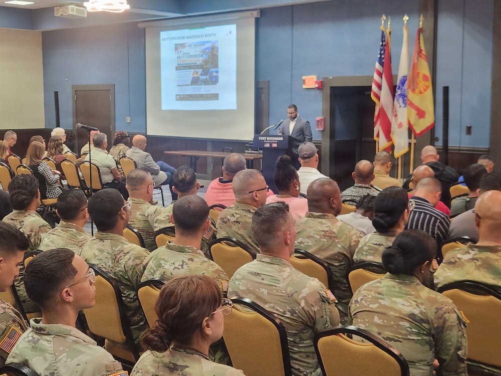 A formal military briefing or presentation is taking place at Fort Buchanan, as indicated by the podium signage. A civilian speaker addresses an audience composed primarily of U.S. Army soldiers in OCP (Operational Camouflage Pattern) uniforms, along with some civilians. The projected slide on screen references 'Antiterrorism Awareness Month' and a '17th Annual Antiterrorism' event.
