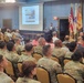 A formal military briefing or presentation is taking place at Fort Buchanan, as indicated by the podium signage. A civilian speaker addresses an audience composed primarily of U.S. Army soldiers in OCP (Operational Camouflage Pattern) uniforms, along with some civilians. The projected slide on screen references 'Antiterrorism Awareness Month' and a '17th Annual Antiterrorism' event.