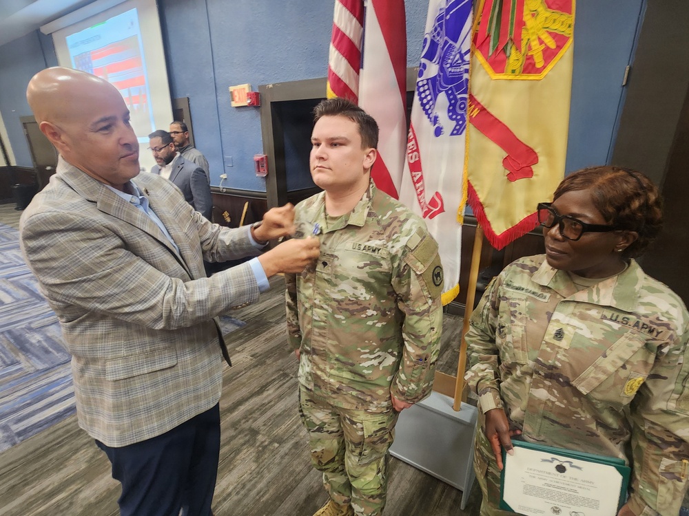 A military ceremony is taking place in what appears to be a conference or meeting room. A civilian man in a blazer is pinning something onto the uniform of a U.S. Army soldier standing at attention. A female U.S. Army NCO stands nearby holding what appears to be a certificate or award document.