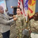 A military ceremony is taking place in what appears to be a conference or meeting room. A civilian man in a blazer is pinning something onto the uniform of a U.S. Army soldier standing at attention. A female U.S. Army NCO stands nearby holding what appears to be a certificate or award document.