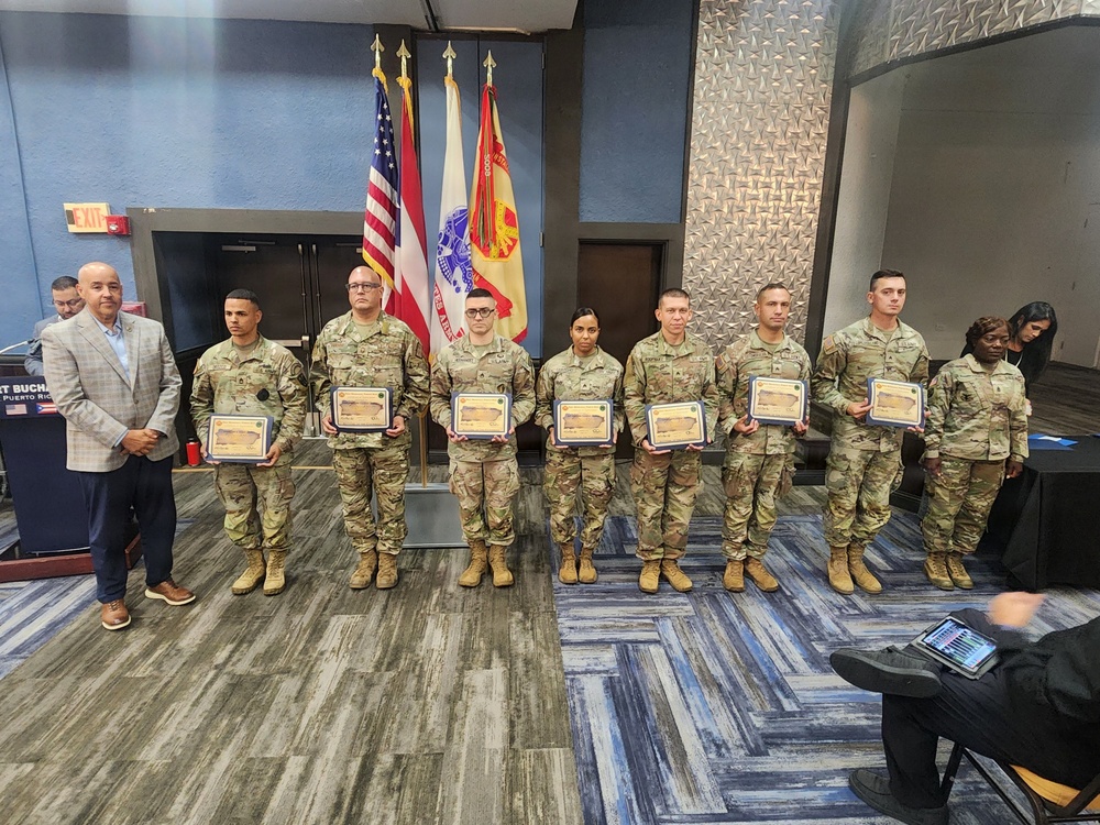A military awards or recognition ceremony featuring seven soldiers in MultiCam/OCP camouflage uniforms holding framed certificates or plaques. They are standing in a line alongside a civilian in a gray blazer, with flags including the U.S. flag, Puerto Rico flag, and U.S. Army flag visible in the background. The setting appears to be an indoor conference or event room.