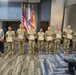 A military awards or recognition ceremony featuring seven soldiers in MultiCam/OCP camouflage uniforms holding framed certificates or plaques. They are standing in a line alongside a civilian in a gray blazer, with flags including the U.S. flag, Puerto Rico flag, and U.S. Army flag visible in the background. The setting appears to be an indoor conference or event room.