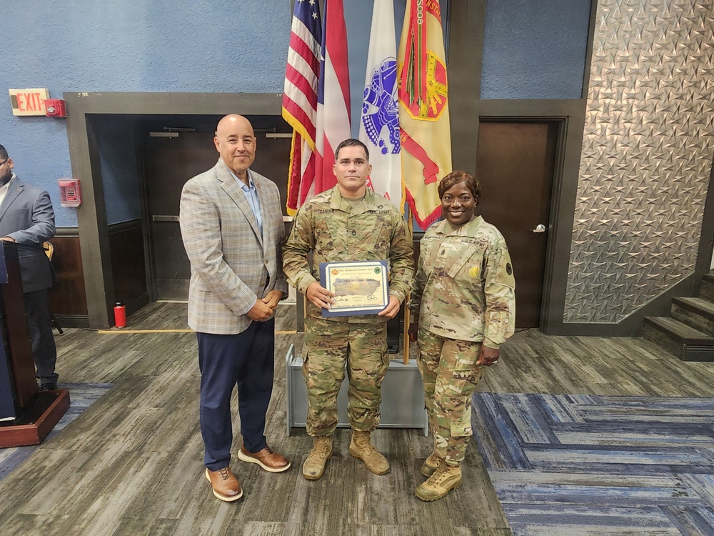 Three individuals pose for a photo at what appears to be a U.S. Army recognition ceremony. A soldier in the center holds a framed certificate or award, flanked by a civilian in a blazer on the left and a female soldier in uniform on the right. Multiple flags, including the U.S. flag and Army-related flags, are visible in the background.