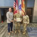 Three individuals pose for a photo at what appears to be a U.S. Army recognition ceremony. A soldier in the center holds a framed certificate or award, flanked by a civilian in a blazer on the left and a female soldier in uniform on the right. Multiple flags, including the U.S. flag and Army-related flags, are visible in the background.