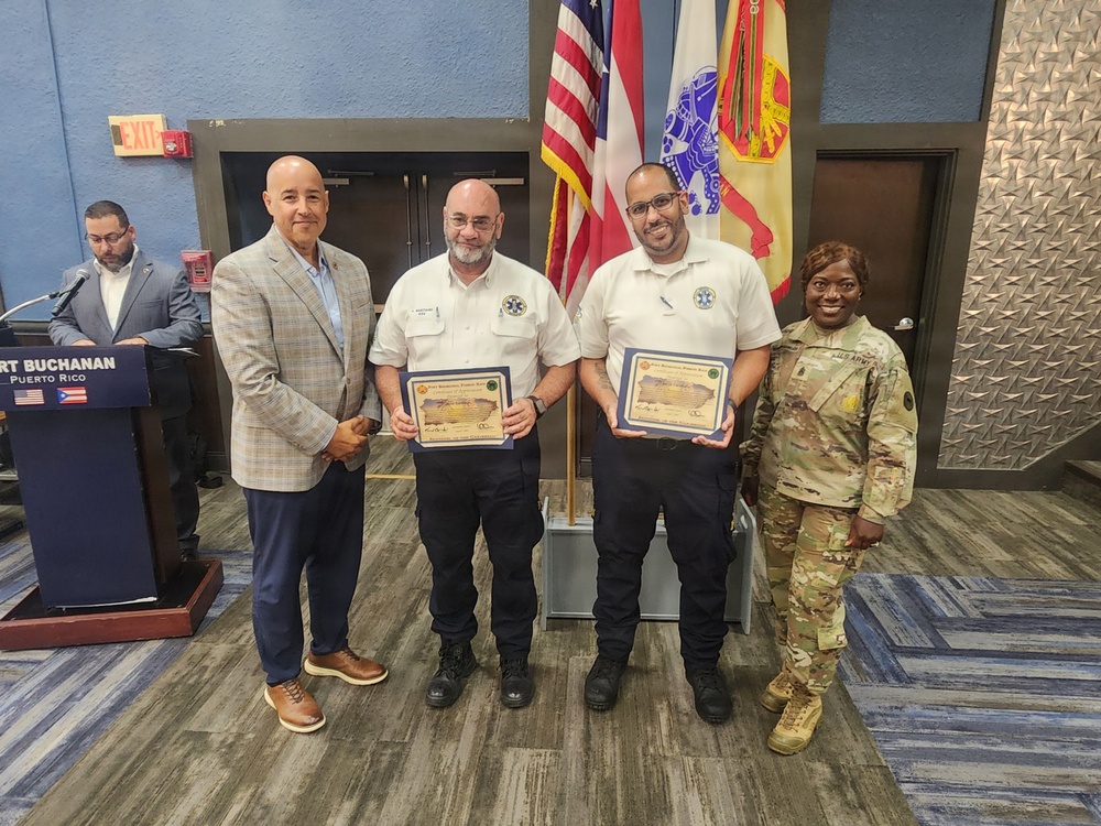 A formal award ceremony is taking place at Fort Buchanan, Puerto Rico, where two individuals in EMS/emergency services uniforms are receiving certificates. They are flanked by a civilian in a plaid blazer and a U.S. Army soldier in OCP camouflage uniform. The setting includes American and Puerto Rican flags along with a U.S. Army flag in the background.