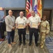 A formal award ceremony is taking place at Fort Buchanan, Puerto Rico, where two individuals in EMS/emergency services uniforms are receiving certificates. They are flanked by a civilian in a plaid blazer and a U.S. Army soldier in OCP camouflage uniform. The setting includes American and Puerto Rican flags along with a U.S. Army flag in the background.
