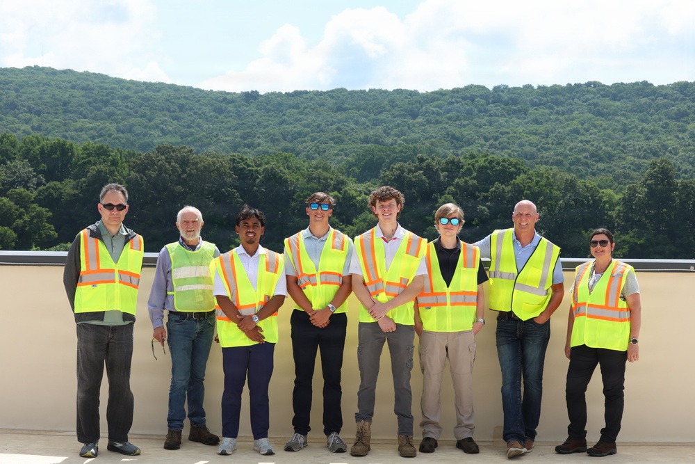 A group of eight people pose together on what appears to be an outdoor terrace or rooftop, with a densely forested hillside visible in the background. Most members of the group are wearing high-visibility safety vests in neon yellow with orange reflective stripes, suggesting a site visit or facility tour. The setting appears to be a warm, sunny summer day.