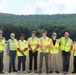 A group of eight people pose together on what appears to be an outdoor terrace or rooftop, with a densely forested hillside visible in the background. Most members of the group are wearing high-visibility safety vests in neon yellow with orange reflective stripes, suggesting a site visit or facility tour. The setting appears to be a warm, sunny summer day.