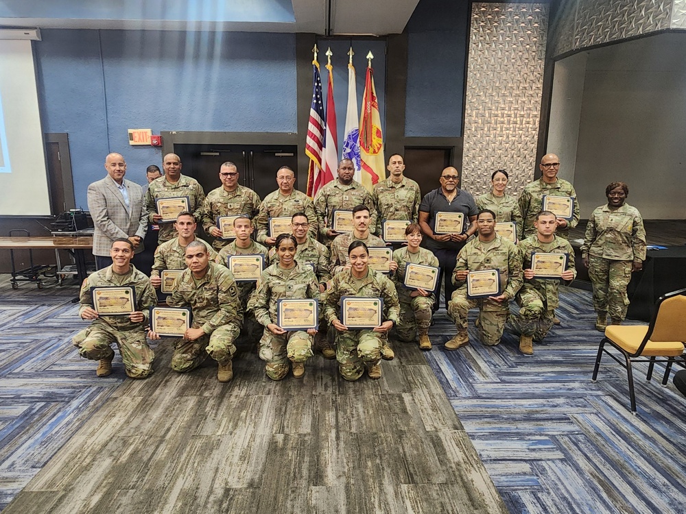 A group of military personnel in camouflage uniforms pose for a formal group photo while holding certificates of achievement or recognition. The ceremony appears to take place in a conference or meeting room, with multiple flags displayed in the background including the U.S. flag and what appears to be the Puerto Rico flag. One civilian in a gray blazer is present among the uniformed service members.