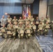 A group of military personnel in camouflage uniforms pose for a formal group photo while holding certificates of achievement or recognition. The ceremony appears to take place in a conference or meeting room, with multiple flags displayed in the background including the U.S. flag and what appears to be the Puerto Rico flag. One civilian in a gray blazer is present among the uniformed service members.