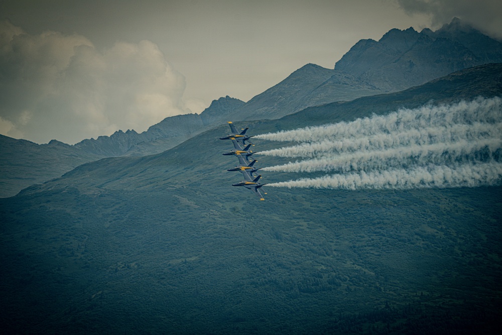 Four military jet aircraft, identifiable by their blue and yellow color scheme as the U.S. Navy Blue Angels flight demonstration squadron, fly in a tight diagonal echelon formation. Each aircraft is emitting white smoke trails as they perform against a backdrop of rugged, forested mountains under an overcast sky. The image captures a precision aerobatic performance, likely at an airshow.