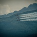 Four military jet aircraft, identifiable by their blue and yellow color scheme as the U.S. Navy Blue Angels flight demonstration squadron, fly in a tight diagonal echelon formation. Each aircraft is emitting white smoke trails as they perform against a backdrop of rugged, forested mountains under an overcast sky. The image captures a precision aerobatic performance, likely at an airshow.