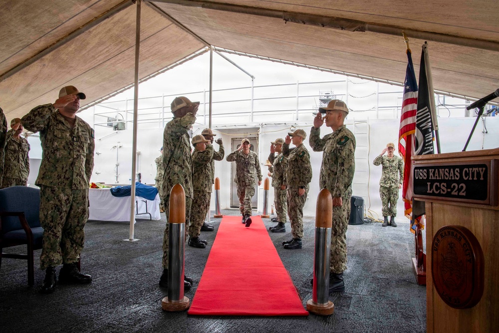 A formal U.S. Navy military ceremony is taking place aboard the USS Kansas City (LCS-22), a Littoral Combat Ship. Personnel in Navy Working Uniform (NWU) camouflage are rendering salutes along a red carpet under a large tent on the ship's deck. A podium bearing the ship's name plate 'USS KANSAS CITY LCS-22' is visible on the right, alongside American and POW/MIA flags.