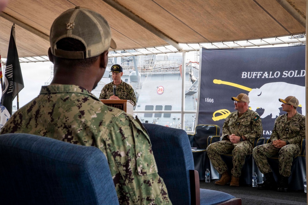 A military ceremony or panel discussion is taking place under a tent or covered outdoor venue, with naval personnel in attendance. A speaker stands at a podium addressing an audience, while two seated military members are positioned near a 'Buffalo Soldiers' banner dated 1866. A naval vessel is visible in the background, suggesting the event is taking place at a naval installation or pier.