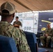 A military ceremony or panel discussion is taking place under a tent or covered outdoor venue, with naval personnel in attendance. A speaker stands at a podium addressing an audience, while two seated military members are positioned near a 'Buffalo Soldiers' banner dated 1866. A naval vessel is visible in the background, suggesting the event is taking place at a naval installation or pier.
