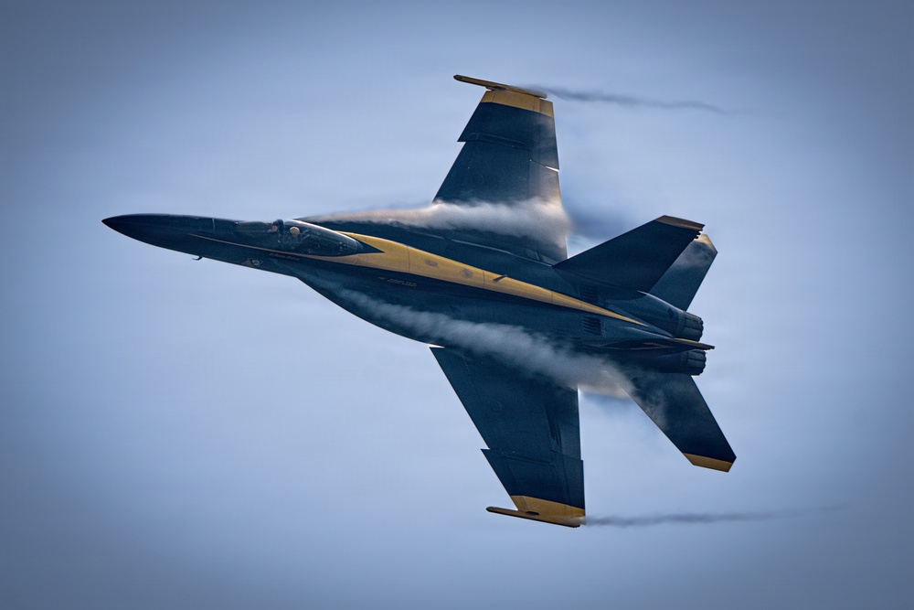 A U.S. Navy Blue Angels F/A-18 Super Hornet is captured in a high-speed, low-altitude pass, viewed from above at a slight angle. The aircraft displays the team's distinctive dark blue and gold livery with visible vapor condensation forming around the fuselage and wings, indicating high-speed flight near the speed of sound. The image is a sharp, professionally composed aerial photograph likely taken at an airshow.