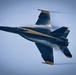 A U.S. Navy Blue Angels F/A-18 Super Hornet is captured in a high-speed, low-altitude pass, viewed from above at a slight angle. The aircraft displays the team's distinctive dark blue and gold livery with visible vapor condensation forming around the fuselage and wings, indicating high-speed flight near the speed of sound. The image is a sharp, professionally composed aerial photograph likely taken at an airshow.