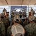 A U.S. Navy ceremony is taking place aboard or dockside near USS Kansas City (LCS-22), featuring a 'Buffalo Soldiers' commemorative event. A speaker addresses an audience of sailors in Navy Working Uniform (NWU) Type III camouflage and tan baseball caps, seated under a white canopy tent. Naval vessels and port infrastructure are visible in the background.