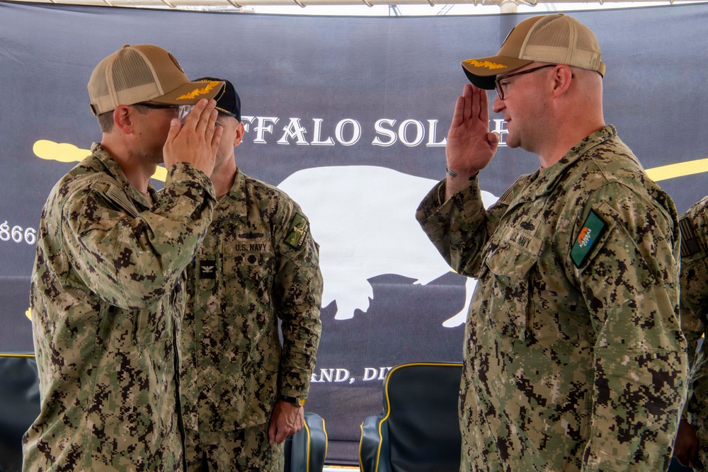 Two U.S. Navy personnel in digital camouflage uniforms are exchanging salutes during what appears to be a formal military ceremony. A third service member stands in the background. The backdrop displays a 'Buffalo Soldier' banner with a buffalo silhouette, suggesting a unit or command ceremony.