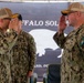 Two U.S. Navy personnel in digital camouflage uniforms are exchanging salutes during what appears to be a formal military ceremony. A third service member stands in the background. The backdrop displays a 'Buffalo Soldier' banner with a buffalo silhouette, suggesting a unit or command ceremony.