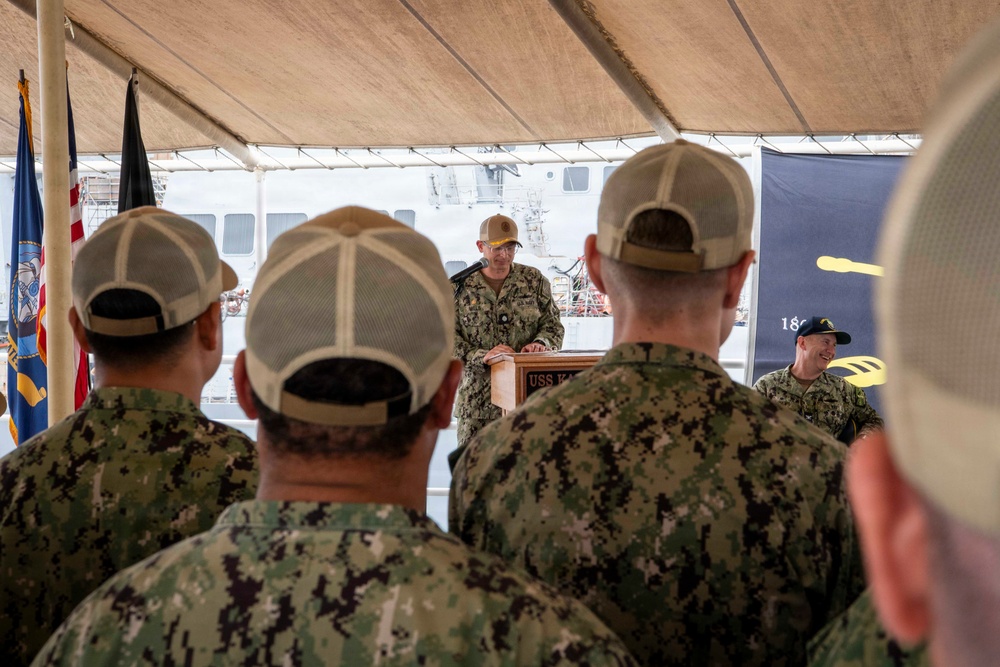 A U.S. Navy ceremony is taking place under a tent, with a uniformed sailor speaking at a podium bearing partial text reading 'USS K...' Attendees in Navy Working Uniform (NWU Type III) camouflage pattern and tan/khaki caps are seated with their backs to the camera. A naval vessel and military flags, including the U.S. flag and what appears to be a Navy flag, are visible in the background.