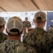 A U.S. Navy ceremony is taking place under a tent, with a uniformed sailor speaking at a podium bearing partial text reading 'USS K...' Attendees in Navy Working Uniform (NWU Type III) camouflage pattern and tan/khaki caps are seated with their backs to the camera. A naval vessel and military flags, including the U.S. flag and what appears to be a Navy flag, are visible in the background.