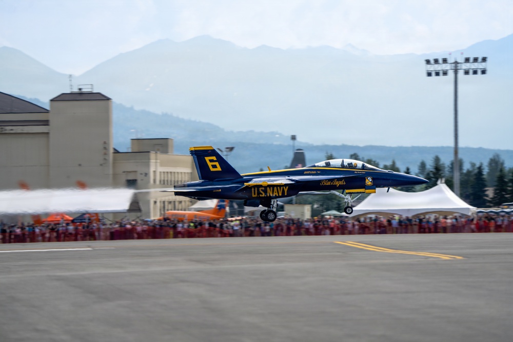 A U.S. Navy Blue Angels F/A-18 Hornet aircraft numbered '6' is captured during a low-altitude pass or takeoff at an airshow. The jet displays the iconic blue and gold color scheme with 'Blue Angels' and 'U.S. NAVY' markings clearly visible. A large crowd of spectators lines the runway behind safety barriers, with mountains and airfield infrastructure visible in the background.