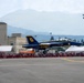 A U.S. Navy Blue Angels F/A-18 Hornet aircraft numbered '6' is captured during a low-altitude pass or takeoff at an airshow. The jet displays the iconic blue and gold color scheme with 'Blue Angels' and 'U.S. NAVY' markings clearly visible. A large crowd of spectators lines the runway behind safety barriers, with mountains and airfield infrastructure visible in the background.