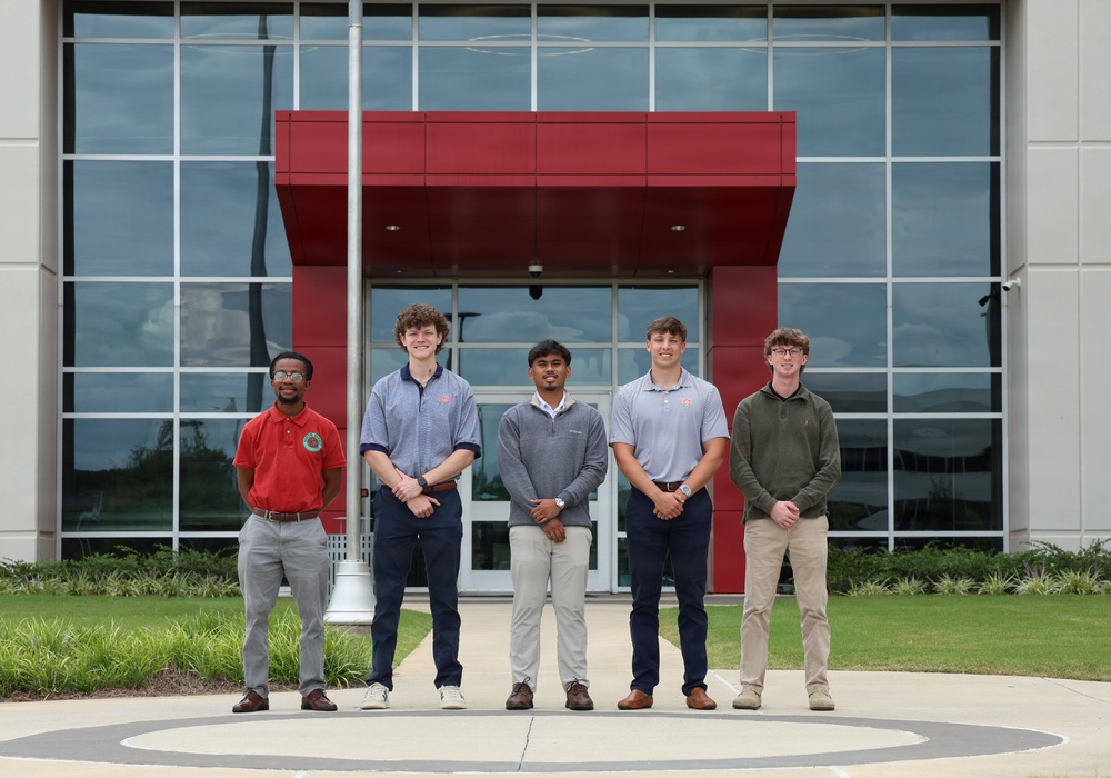 Five young men are posed in a group photo in front of a modern commercial or institutional building featuring red and glass architectural elements. They are standing on a decorative circular pavement design, dressed in business-casual attire, suggesting a professional or academic occasion. The setting and their coordinated posture indicate this may be an internship, scholarship, or recognition photo.