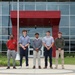 Five young men are posed in a group photo in front of a modern commercial or institutional building featuring red and glass architectural elements. They are standing on a decorative circular pavement design, dressed in business-casual attire, suggesting a professional or academic occasion. The setting and their coordinated posture indicate this may be an internship, scholarship, or recognition photo.