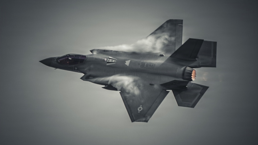 A fifth-generation stealth fighter jet, consistent with an F-35 Lightning II, is captured in a banked maneuver from a top-down perspective. Condensation vapor trails are visibly forming over the fuselage and wings due to high-speed aerodynamic pressure changes. The image is rendered in a desaturated, near-monochromatic tone with the engine afterburner providing a subtle warm glow.