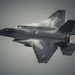 A fifth-generation stealth fighter jet, consistent with an F-35 Lightning II, is captured in a banked maneuver from a top-down perspective. Condensation vapor trails are visibly forming over the fuselage and wings due to high-speed aerodynamic pressure changes. The image is rendered in a desaturated, near-monochromatic tone with the engine afterburner providing a subtle warm glow.