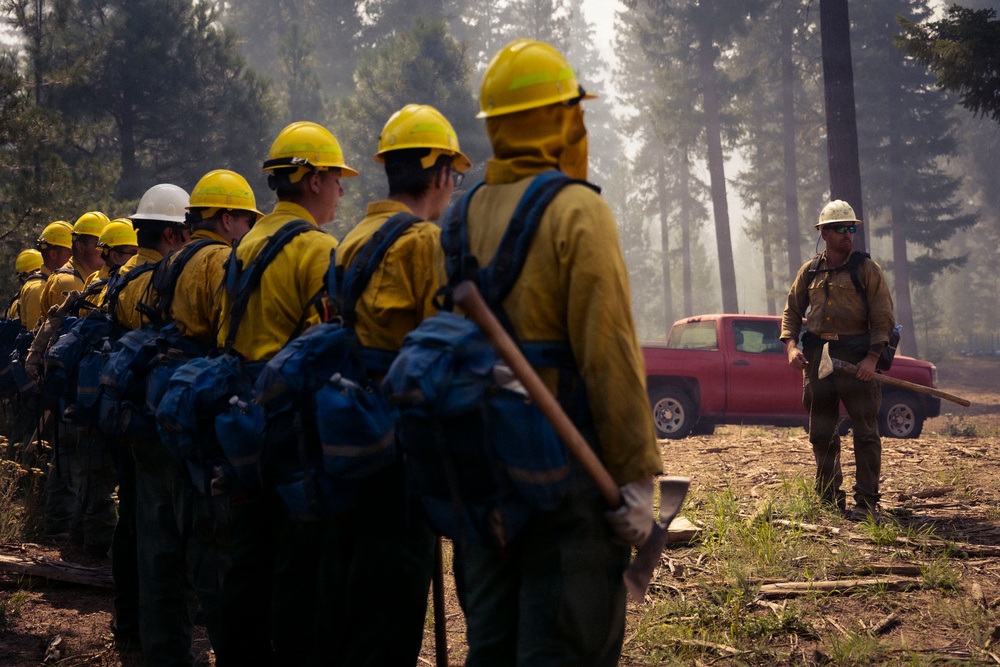 A crew of wildland firefighters in yellow Nomex shirts and hard hats stands in formation in a smoky forest clearing, facing a supervisor or crew boss who is addressing them. The crew is fully equipped with blue backpacks and hand tools including axes and pulaskis. The scene suggests a pre-deployment briefing or morning lineup at a wildfire incident.