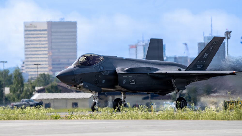 A dark gray F-35A Lightning II stealth fighter jet is taxiing on a runway, with tail code 'AK' and serial number 5534 visible, indicating assignment to an Alaska-based unit. The aircraft is photographed in an urban airfield environment with a city skyline visible in the background. The image captures the aircraft at ground level with landing gear deployed.