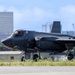 A dark gray F-35A Lightning II stealth fighter jet is taxiing on a runway, with tail code 'AK' and serial number 5534 visible, indicating assignment to an Alaska-based unit. The aircraft is photographed in an urban airfield environment with a city skyline visible in the background. The image captures the aircraft at ground level with landing gear deployed.