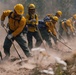 A crew of wildland firefighters in yellow Nomex gear and hard hats work in a coordinated line, digging and scraping the ground with hand tools to construct a fire line. Dust fills the air as the crew works in unison across a smoky, disturbed landscape. The image captures the intense physical labor involved in wildfire suppression operations.