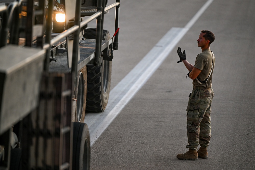 A military service member in camouflage uniform is directing a large military vehicle on what appears to be a tarmac or airfield. The soldier is wearing tactical gloves and using hand signals to guide the vehicle. The image is taken during low-light conditions, likely at dusk or dawn.