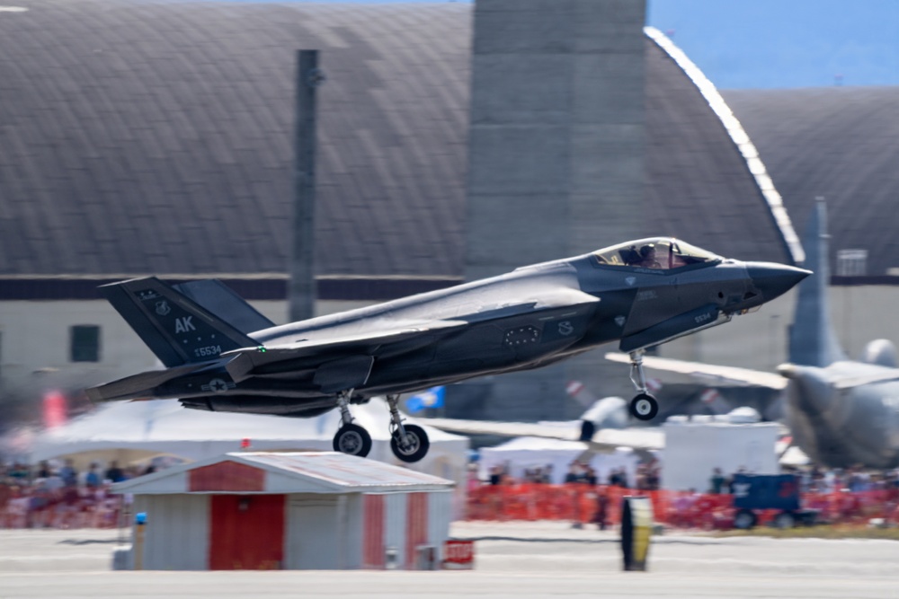 An F-35A Lightning II fighter jet bearing 'AK' tail code and serial number 15534 is captured during takeoff or low-altitude flight at what appears to be an airshow. The aircraft is in a dark gray stealth finish with landing gear still deployed. A crowd of spectators and military hangars are visible in the background.